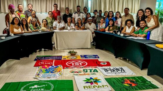 Um grupo de 40 pessoas sentados numa mesa olhando para a câmera e bandeiras dos movimentos ao chão compõe o cenário